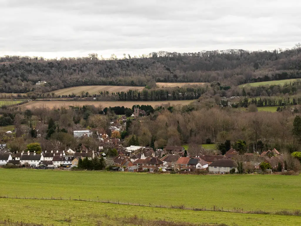 Looking down the grass hill to a village bellow surrounded by trees, and a hill rising up the other side.