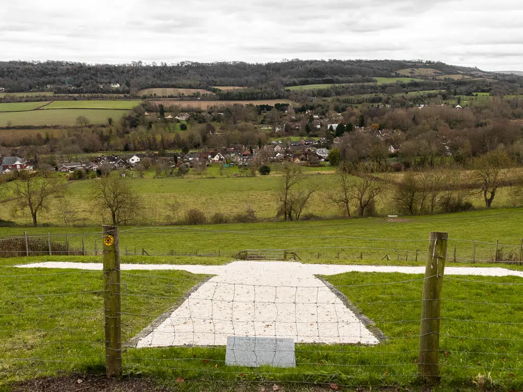 Looking down the hill over the white chalk crosstown a village below surrounded by trees.