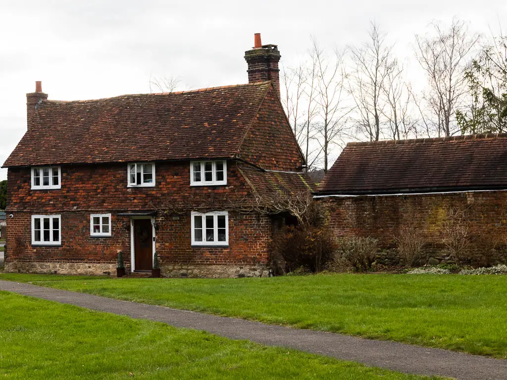 A path cutting diagonally through a green with an old cottage on the other side, with white framed windows.