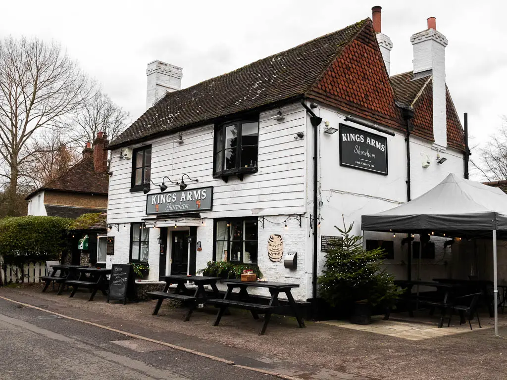 A white walled pub with a black sign on it saying 'Kings arms shoreham'. There are black picnic benches in front of it, and a black gazebo to the right. 