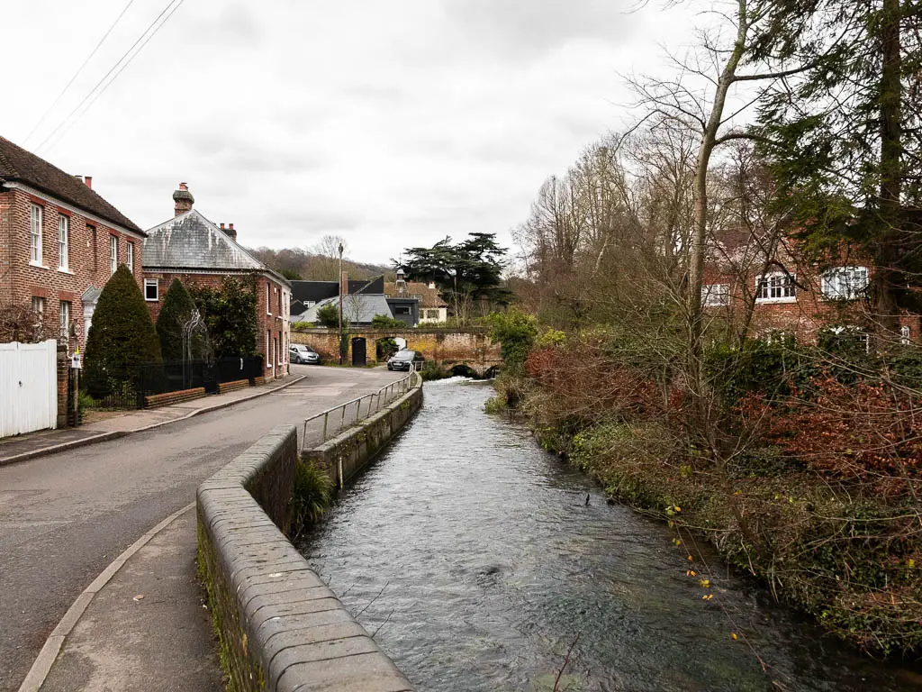 A river on the right, and road on the left. The right side of the river is lined with bushes and trees, the left side of the road is lined with houses. 