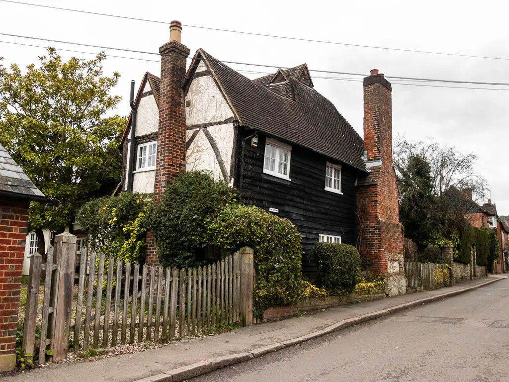 A black timber framed old cottage with a red brick chimney up the side. There are hedges around the left side of the cottage, and a wooden fence to the left. The cottage sits next to the road.