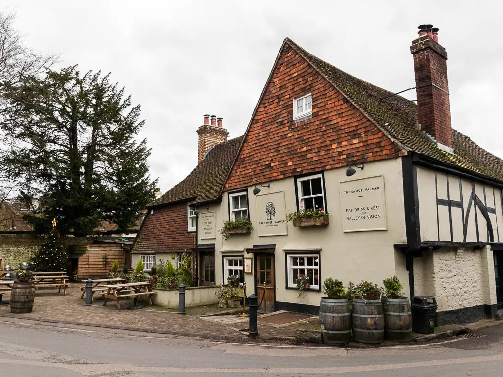 A pub on a road corner, with three barrels with plants next to it. There are wooden picnic benches to the left. There is a chimney coming out of the roof.