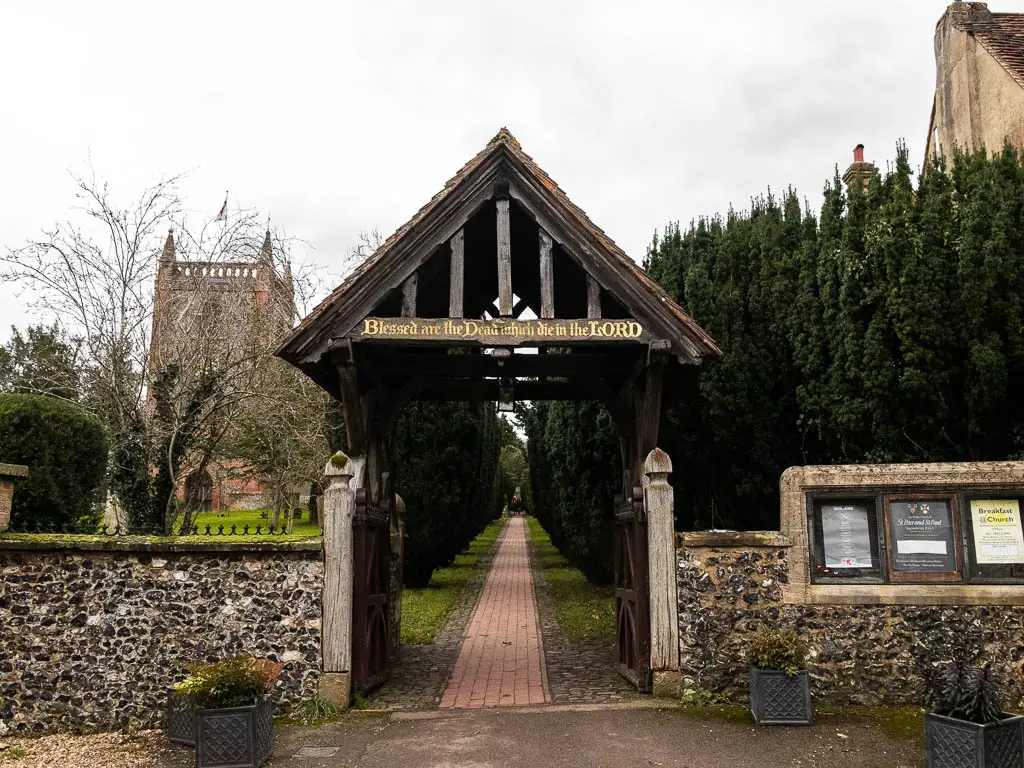 Looking towards a path through the stone wall, leading under an A framed arch. There is information boards on the right, and part of the church visible to the left.