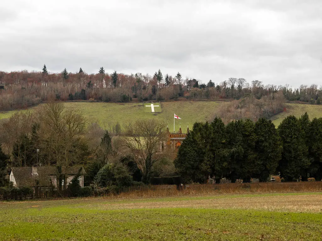 Looking across a large grass field towards a mass of trees and a few hoses and a church poking through. There is a hill past the trees with a white cross on it. There are trees at the top of the hill.