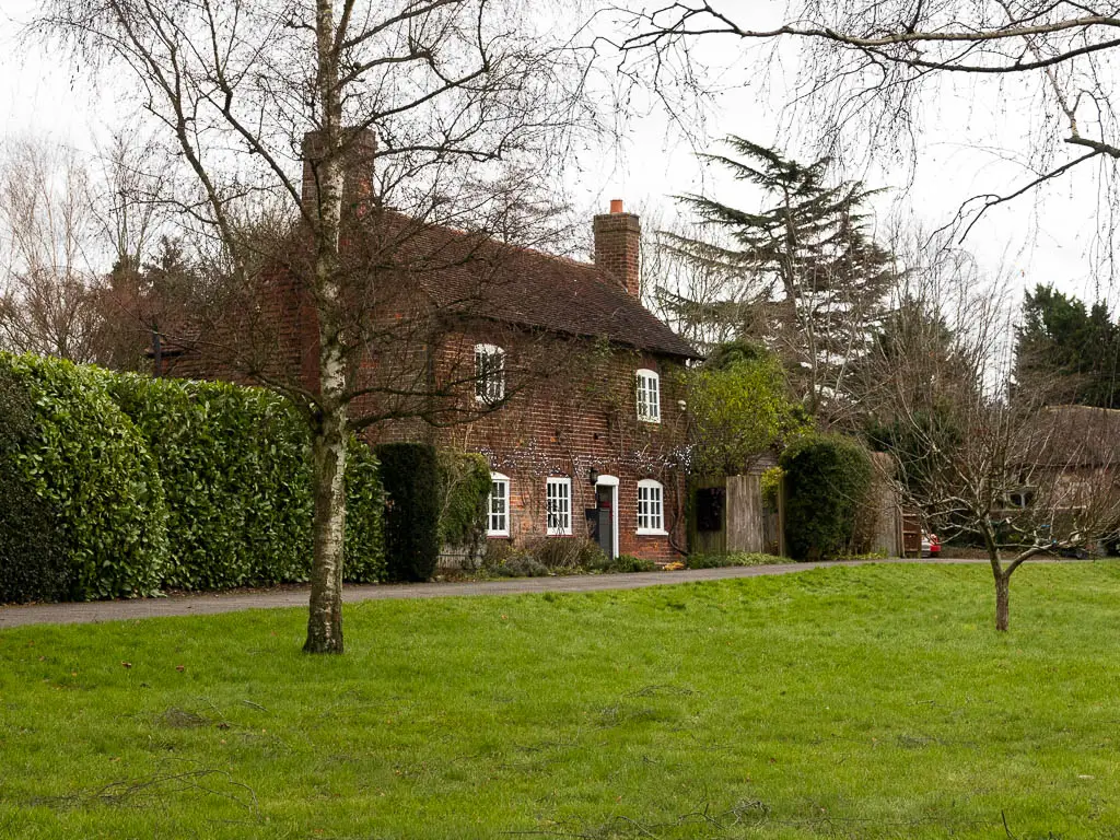 Looking across a neatly cut green to an old cottage with white framed windows. There is a neatly cut hedge to the left of the cottage. There are a couple of leafless trees on the green.