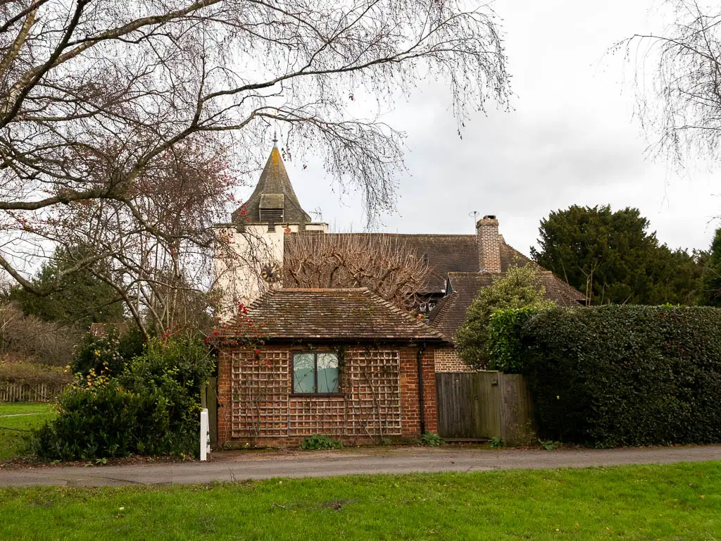 A strip of green with a path running from left to right, and cottages on the other side of the path. The cottages are surrounded bye hedges and bushes. There are leafless tree branches hanging into frame from the left.