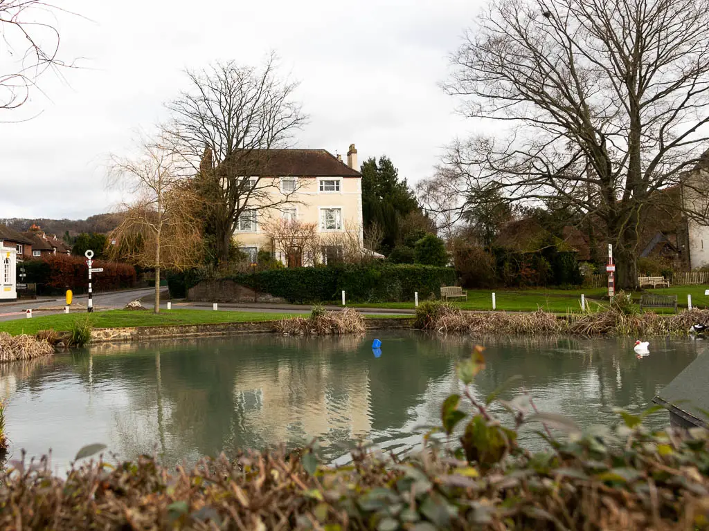 Looking across the otford pond, on the way to Shoreham. There is a white walled house on the other side. there are a couple of large leafless trees on the other side.