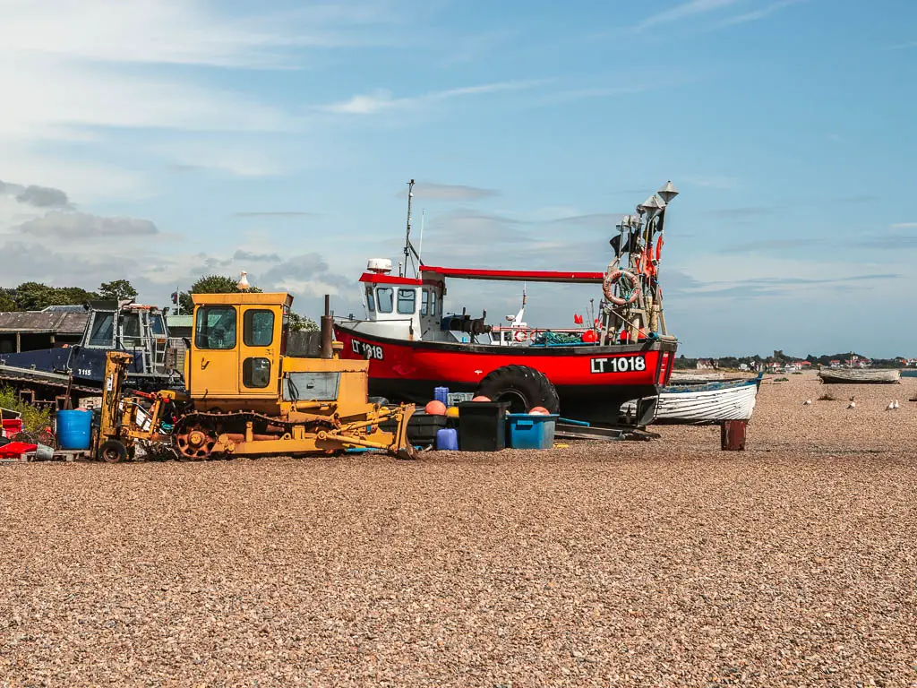 A red fishing boat, yellow plough, and other stinging items sitting on the shingle beach in Aldeburgh.