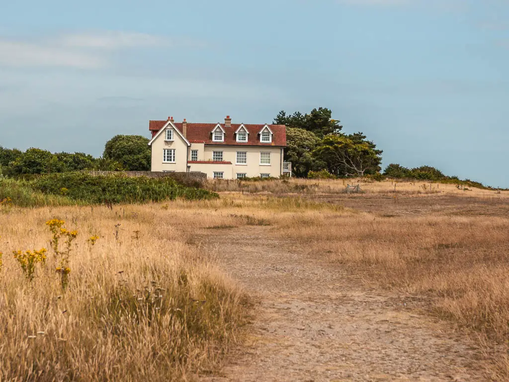 A wide trail through the meadow, leading towards a cream walled house with res roof, on the walk from Snape to Dunwich.