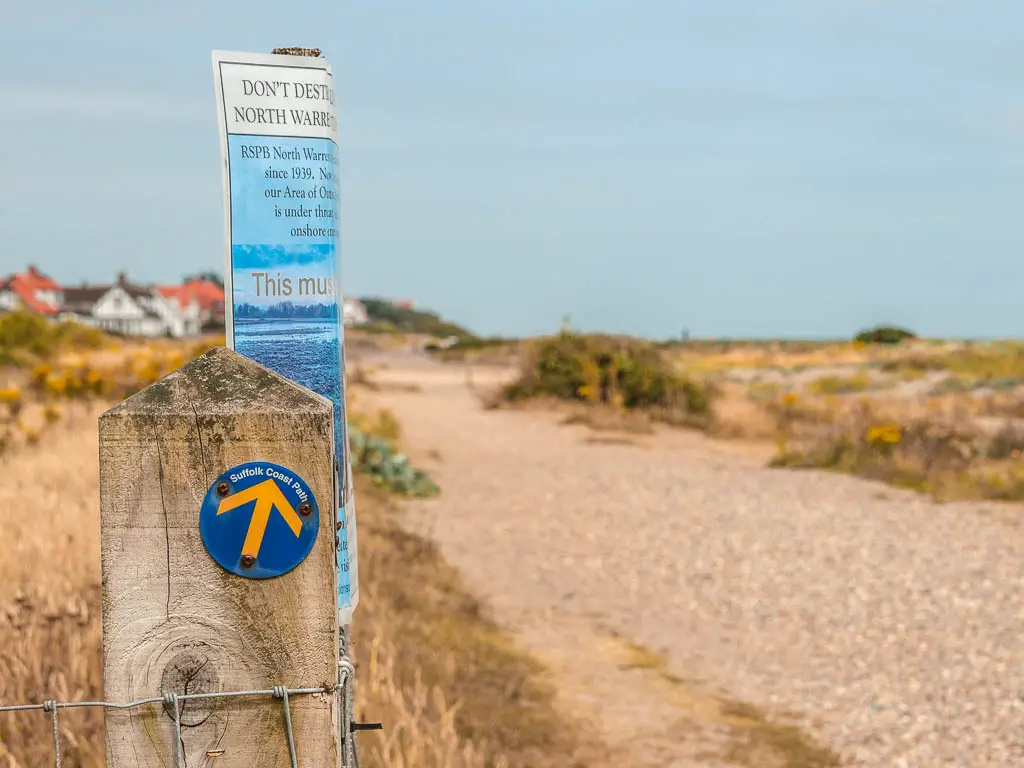 A blue Suffolk coast path disc on a wooden post, on the walk from Snape to Dunwich. It has a yellow arrow pointing ahead along the path.