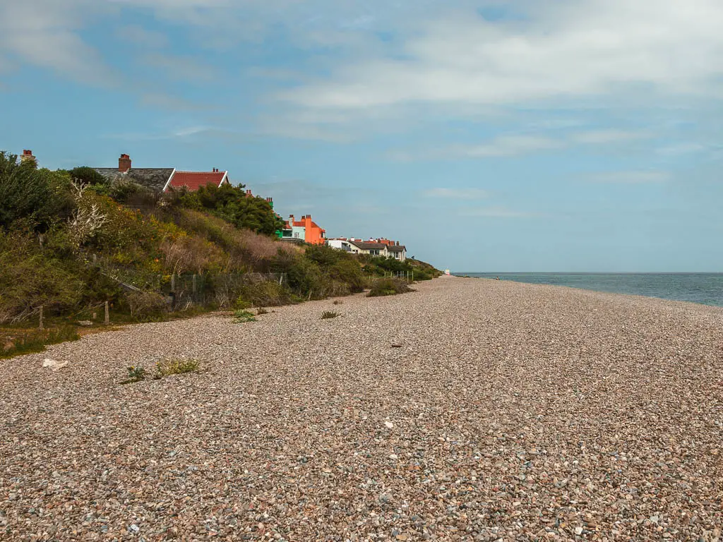 A shingle beach leading ahead, with a green bush and grass covered small hill to the left, and blue sea to the right.