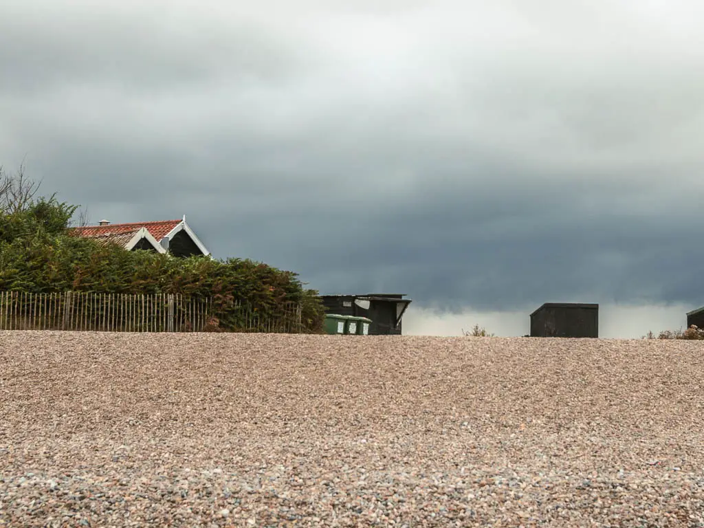 Looking up the shingle beach to some black huts next to a wooden fence. There are green bushes on the other side of the fence, with a rooftop poking out the top.