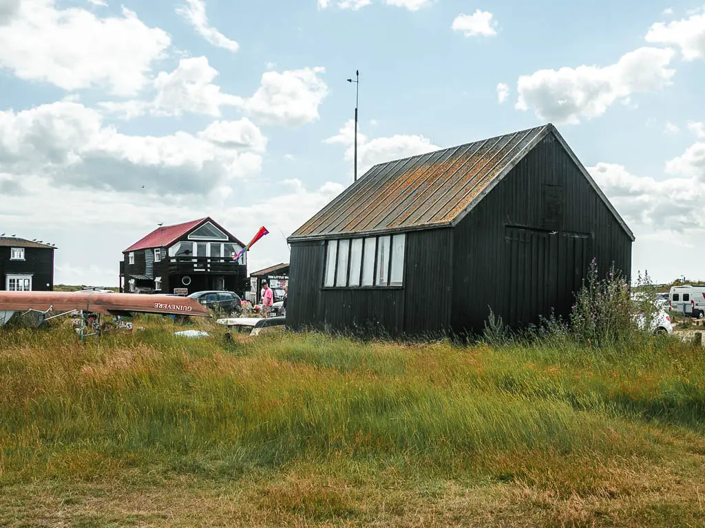 A black wooden hut sitting in the tall grass, with an upturned boat on the left and more black huts ahead in the distance. 