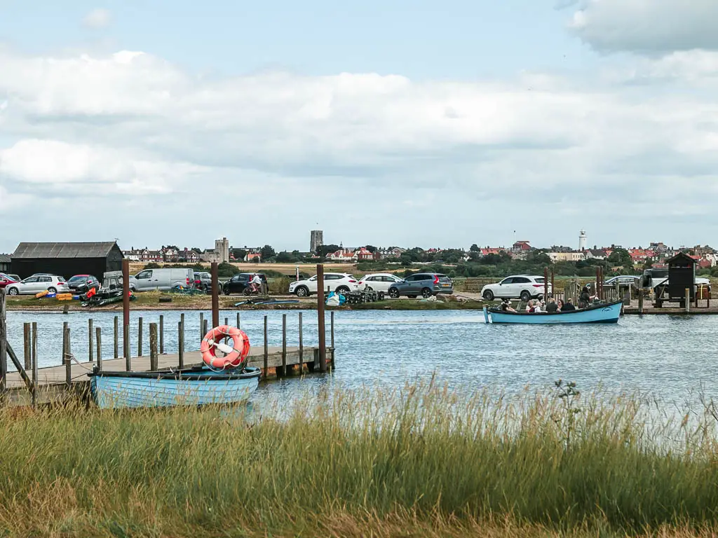 Looking over the tall green grass to a river, with a pier leading into it, on the walk from Dunwich to Kessingland. There is a blue row boat moored to this side of the river, and a blue boat filled with people on the other side.