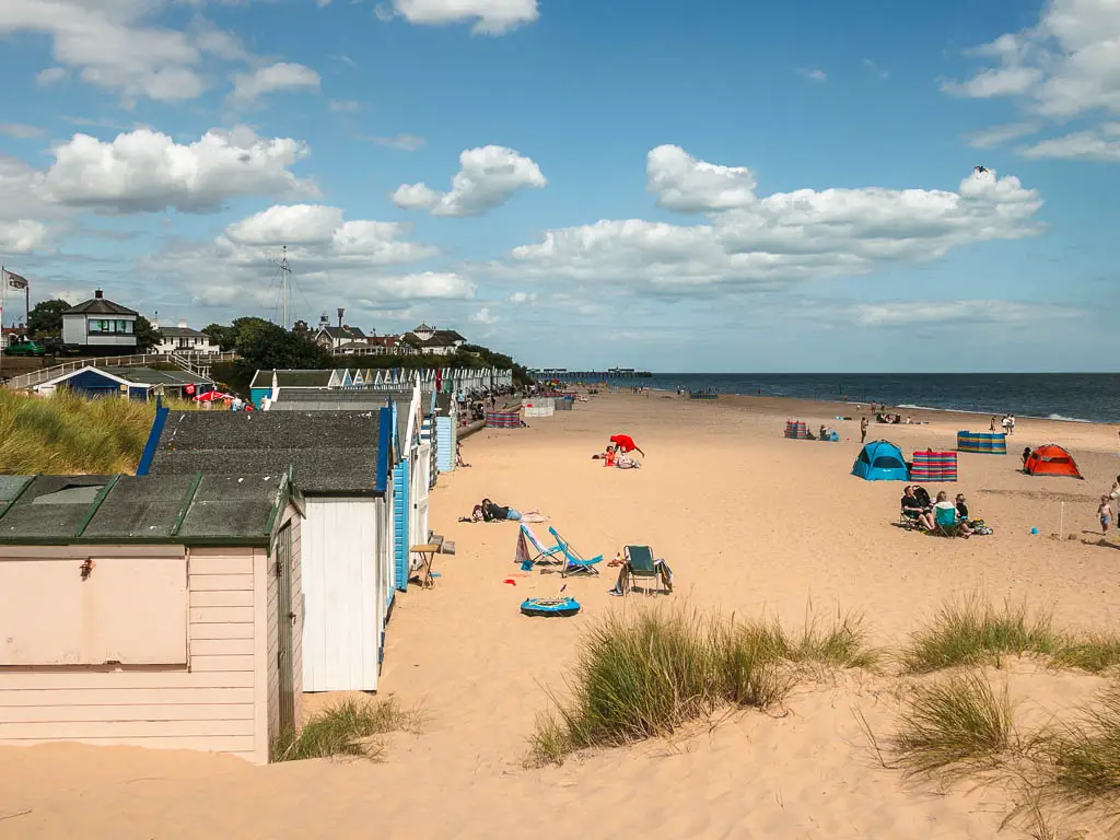 Looking down and along a large sandy beach, with a few people and dec chairs on it, and beach huts lining the left side.