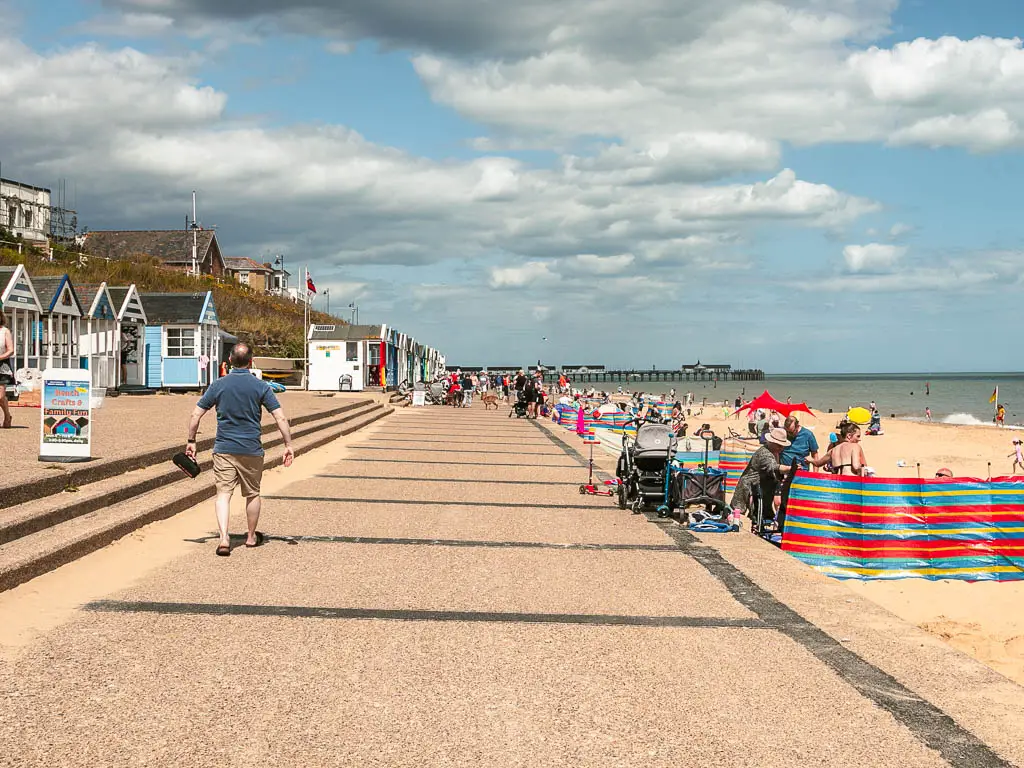 A wide promenade leading straight ahead, with a sandy beach filled with people to the right. There is a man walking along the promenade. 