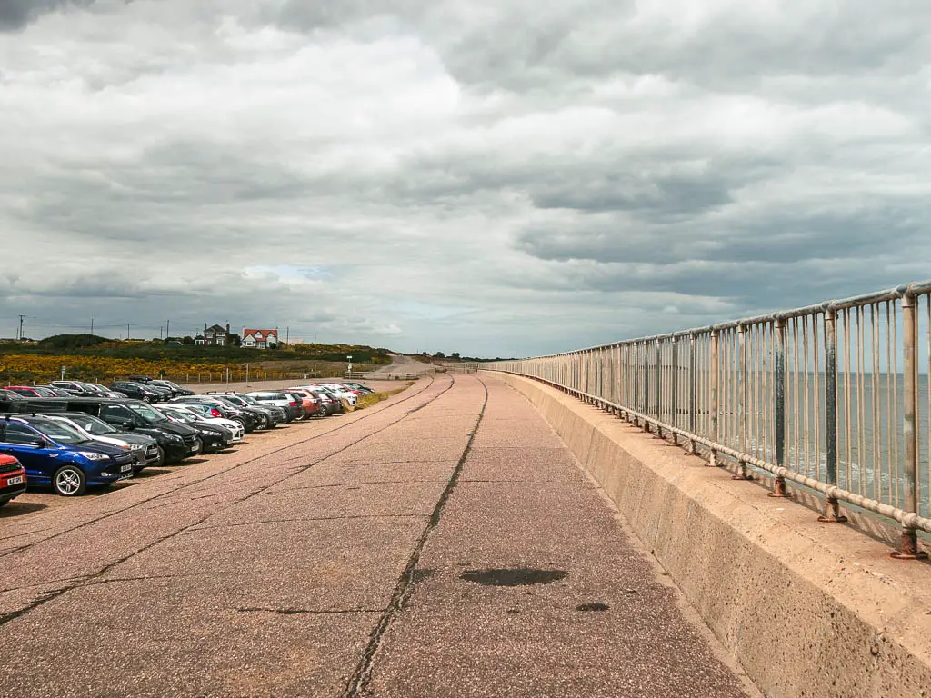 A long straight walkway with metal railings to the right, and a mass of cars parked in the car park to the left.