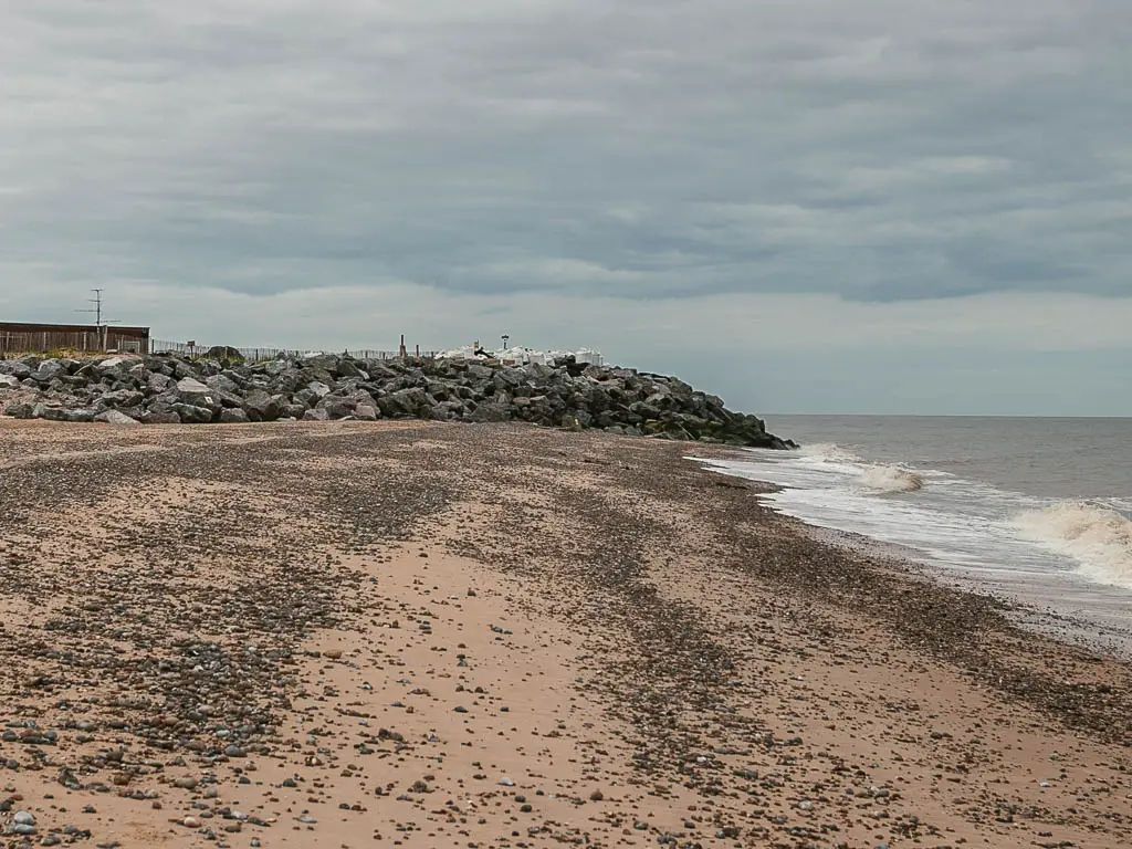 A sandy beach with pebbles leading ahead to a mass of rocks, with the rough North Sea to the right.