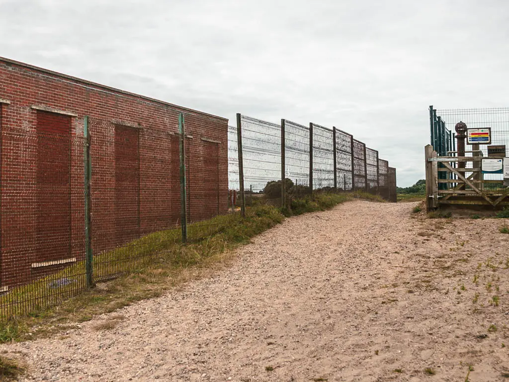 A sandy path with a tall metal fence on the right, with a brick block building on the other side.