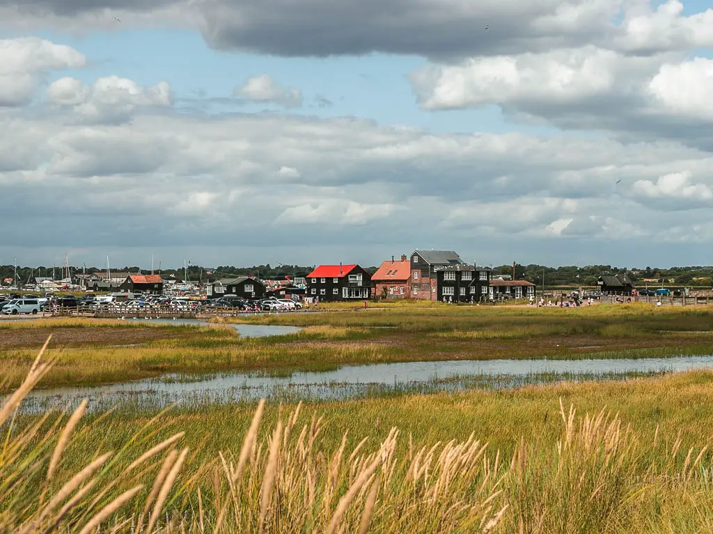 Looking across the marshland to a mass of civilisation on the other side, with timber hoses, cars, and people.