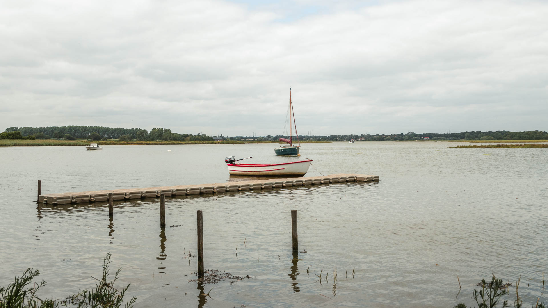 A small white motor boat sitting in the middle of the river, with a wooden walkway next to it, on the walk from Orford to Snape. There is a sailboat behind it, with its sails down.