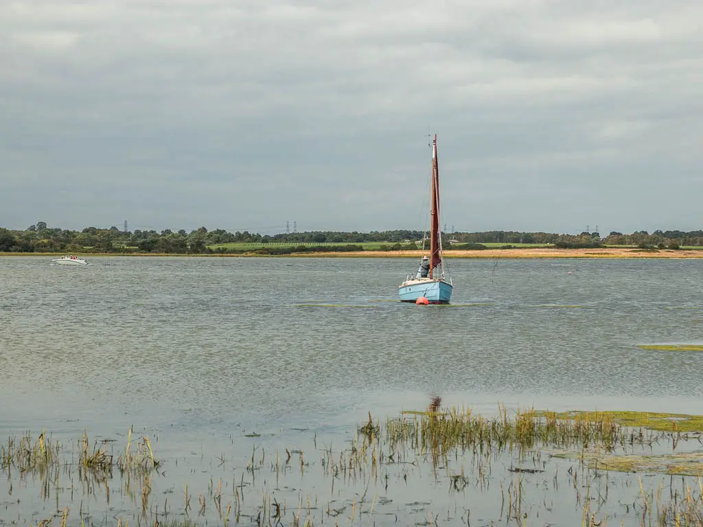 A blue sailboat sitting just off centre in the river, on the walk between Orford and Snape. There are ripples in the river.