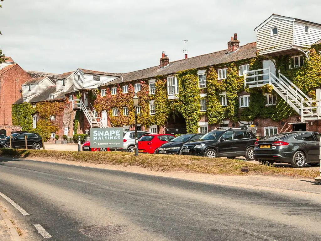 Looking across the road to a large budding covered in ivy in Snape. There are cars parked outside, and a sign that says 'Snape Maltings'.